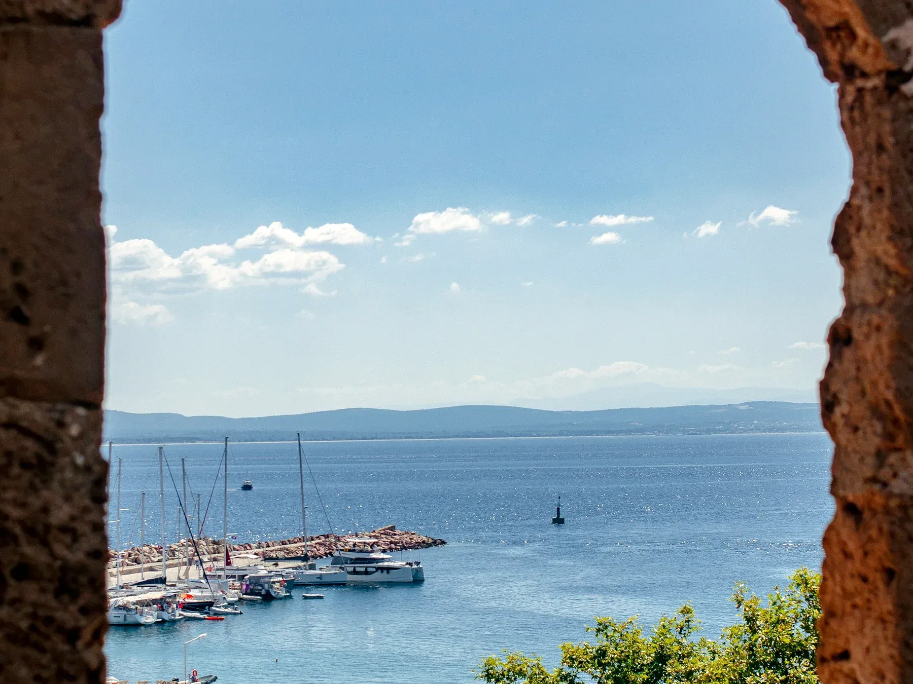 A view through a rough stone window opening onto a small marina of moored sailboats and yachts on the blue Adriatic sea, with hills along the far coastline.