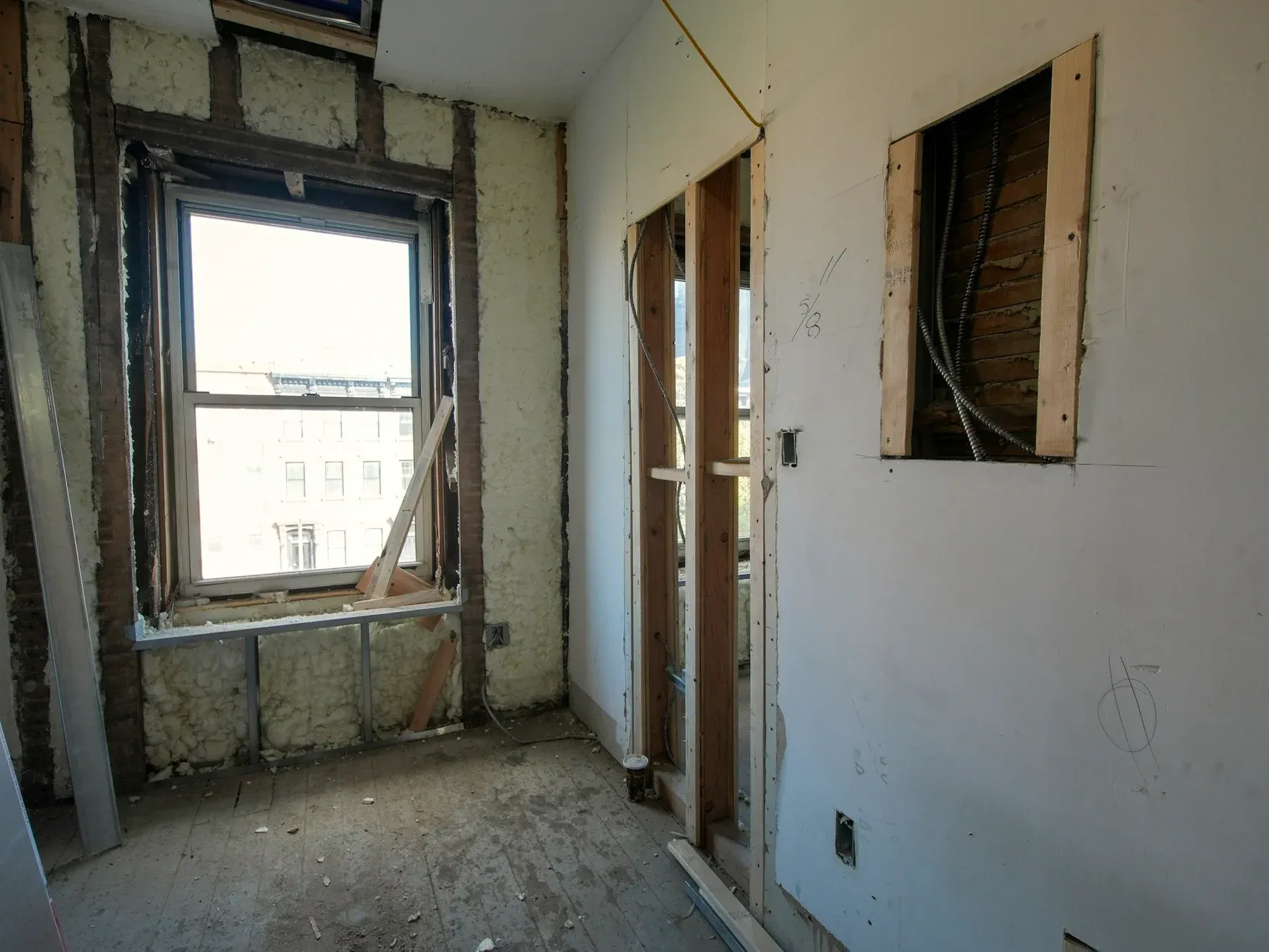 An interior room undergoing renovation, with exposed timber stud framing, stripped drywall, spray-foam insulation around a window and exposed electrical wiring.