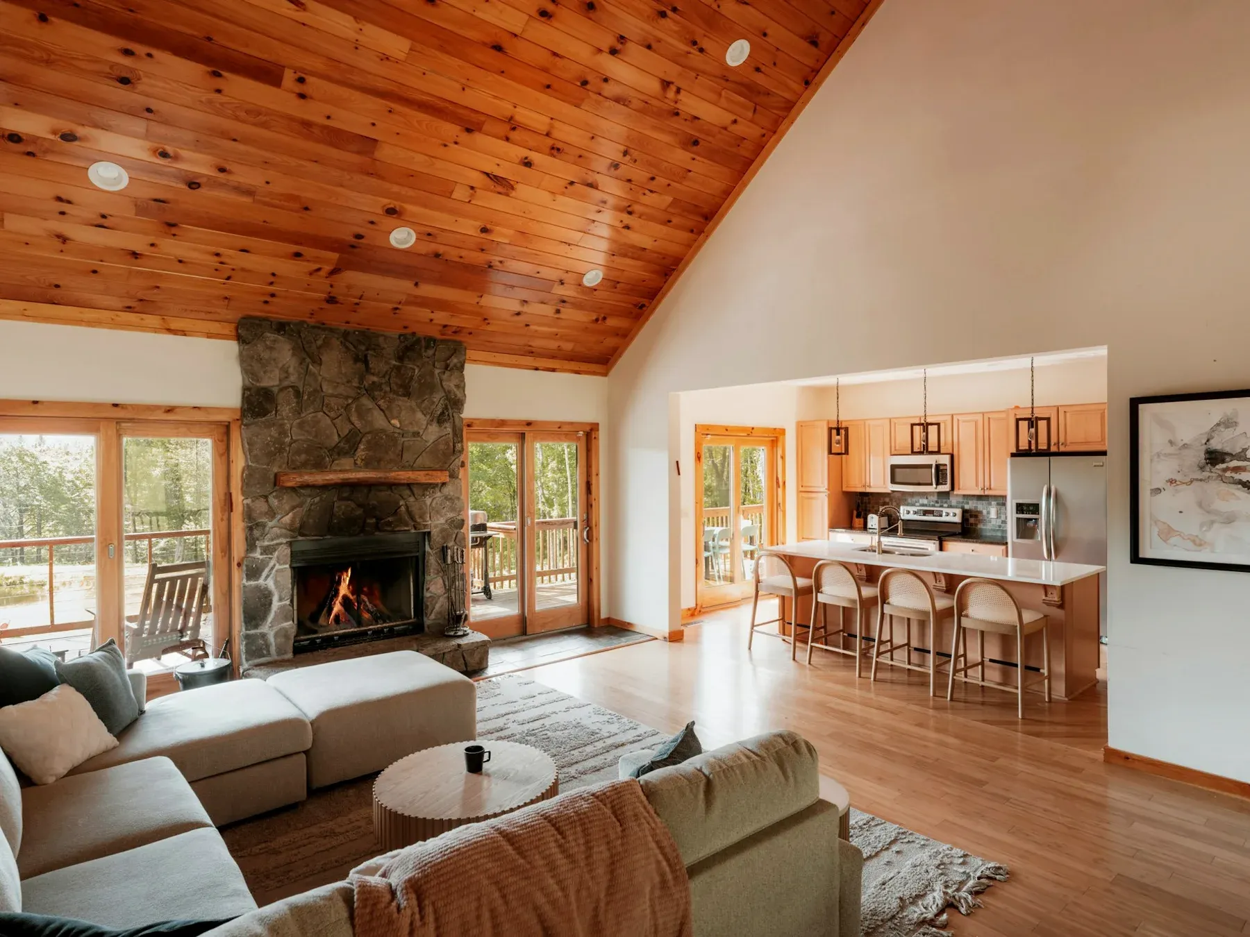 A calm living room with a stone fireplace, timber ceiling and wide windows opening onto surrounding greenery.