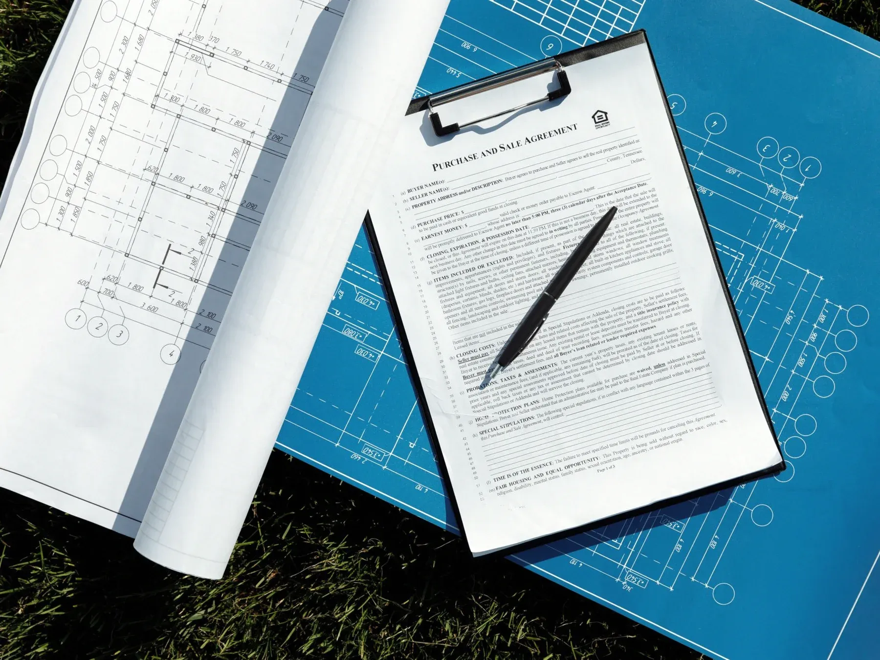 A printed purchase-and-sale agreement on a black clipboard with a pen, resting on rolled paper drawings and blue architectural blueprints.