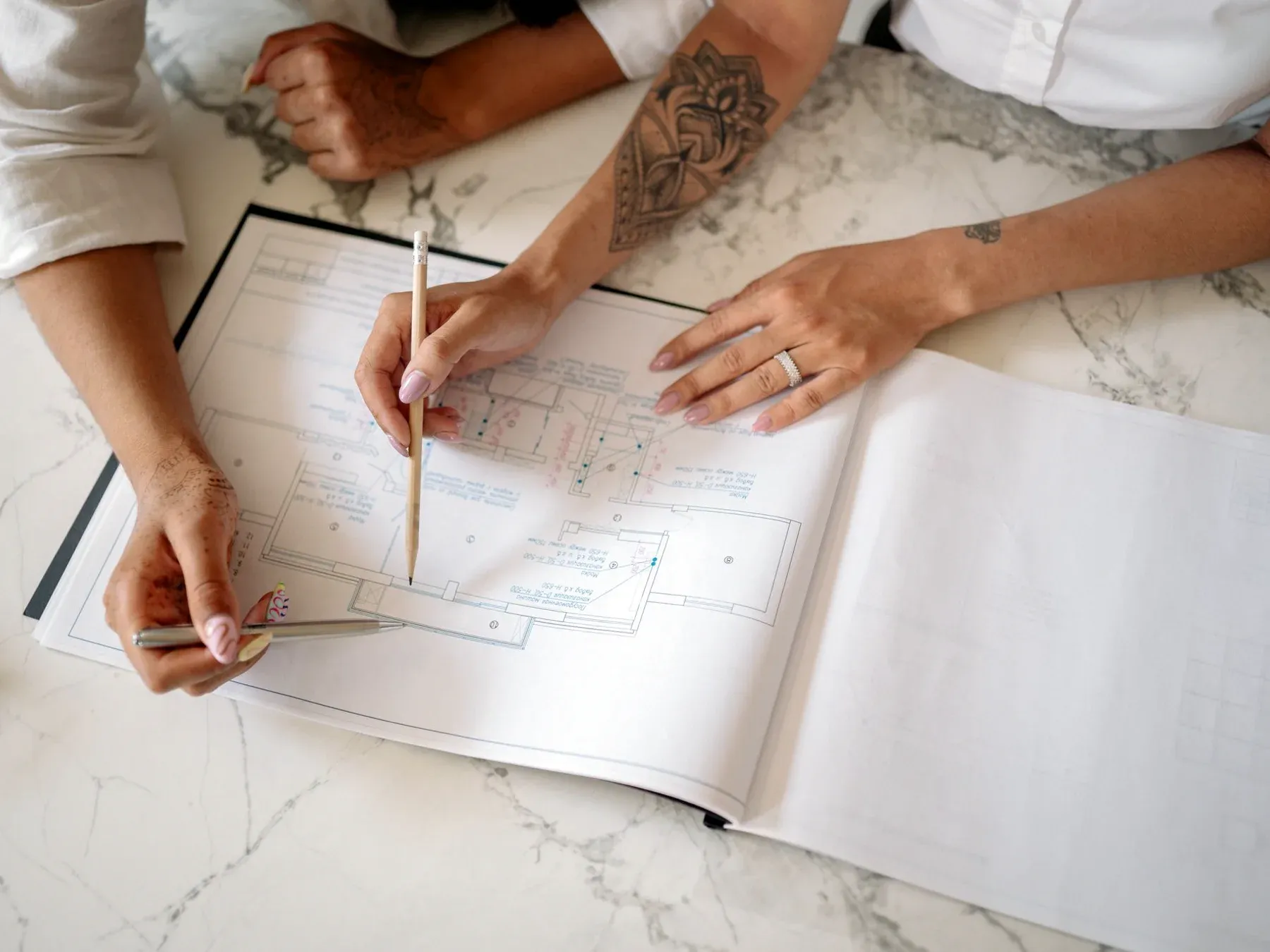An overhead view of two people at a marble table studying a printed architectural floor plan, one marking the drawing with a pencil.