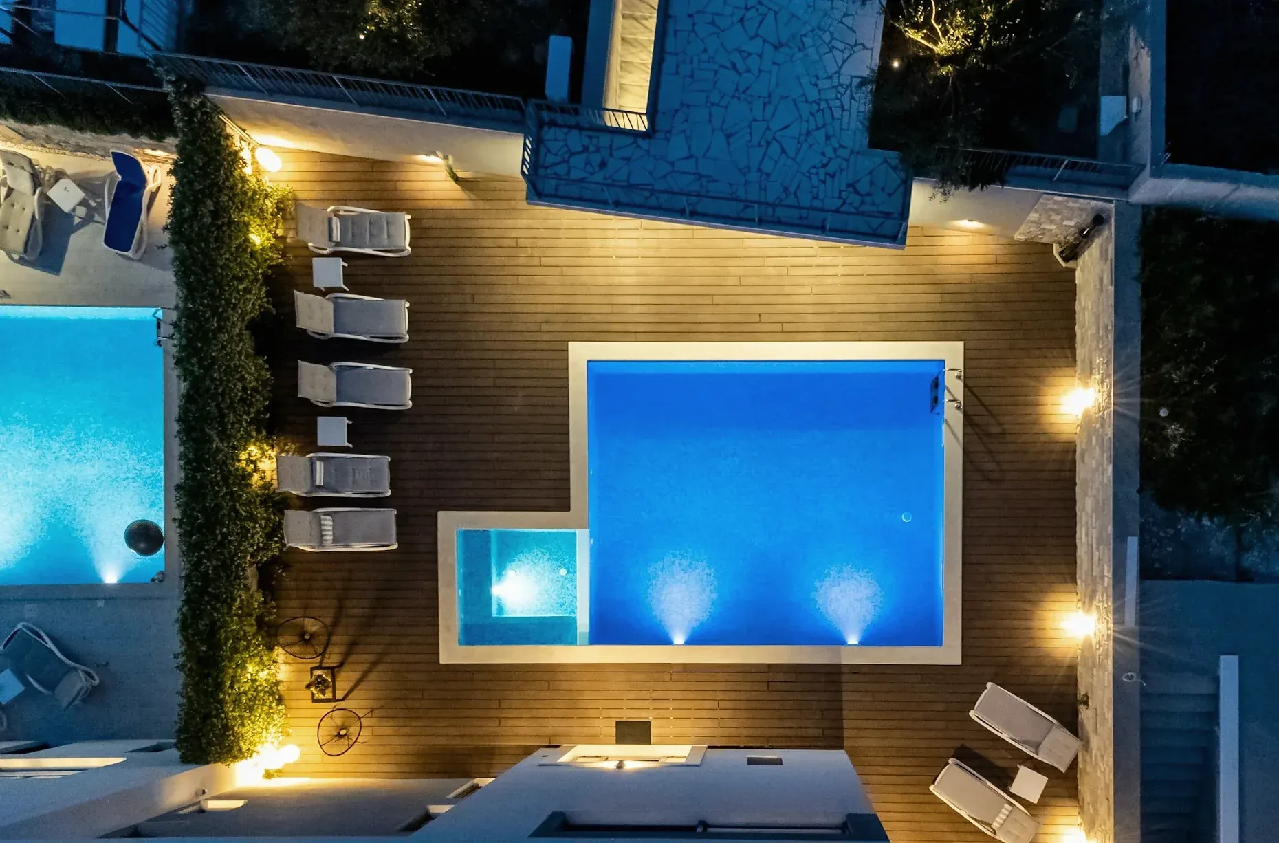 An overhead night view of a Croatian villa's lit pool, timber deck and sun loungers.