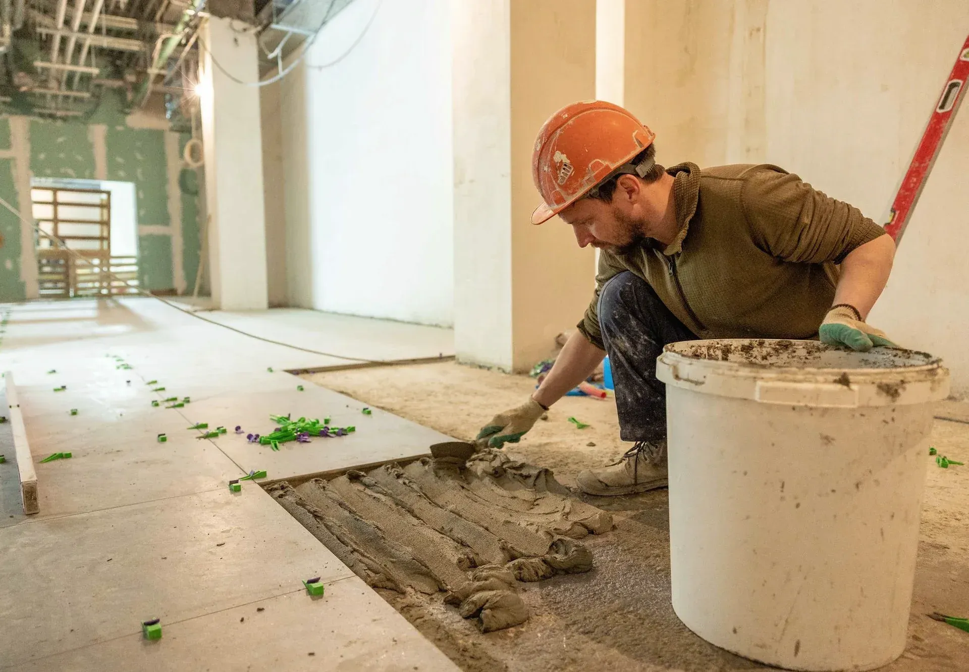 A worker in a hard hat spreading adhesive to lay floor tiles on a renovation site.