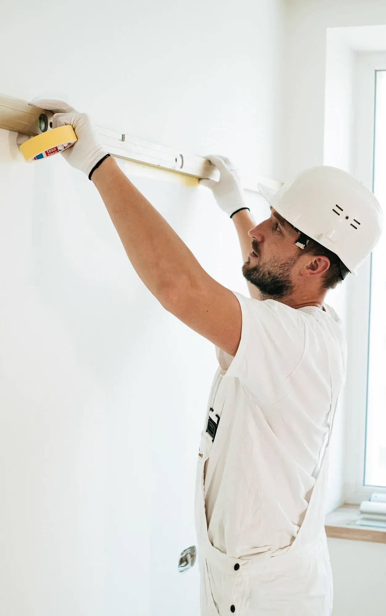 A worker in white overalls and a hard hat holding a spirit level against a wall to check it is level during building work.