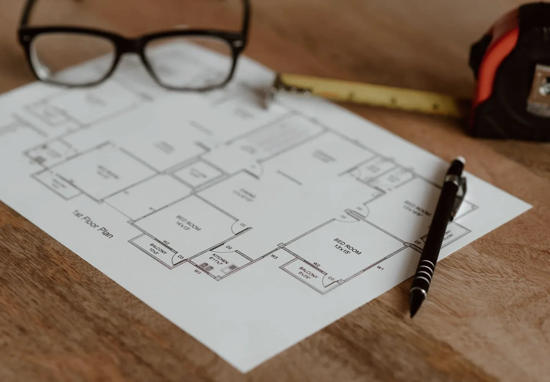 A printed floor plan on a desk with a pen, folding ruler, tape measure and reading glasses.
