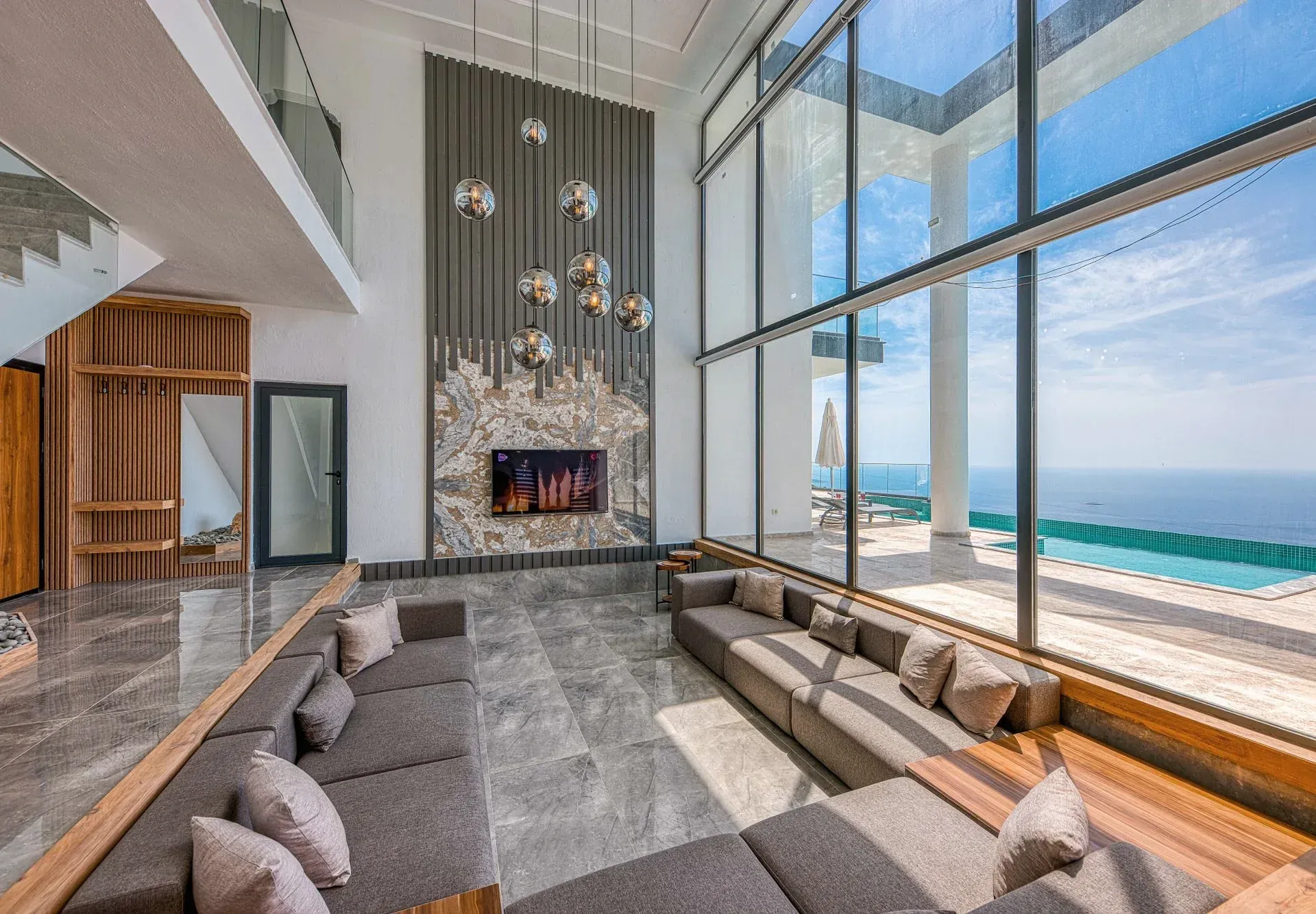 A double-height seaside villa living room with floor-to-ceiling windows framing the sea and an infinity-pool terrace beyond.